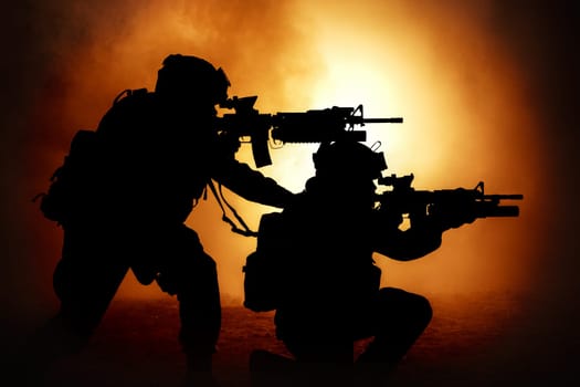 Silhouettes of two army soldiers, U.S. marines team in action, surrounded fire and smoke, shooting with assault rifle and machine gun, attacking enemy with suppressive gunfire during offensive mission