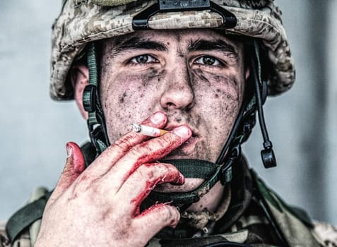 Close up portrait of wounded soldier, U.S. marine with dirty face, holding cigarette with soiled in blood fingers and smoking. Military medic resting after field operation. Traumatic war experiences