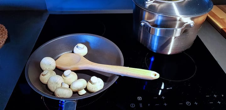 A silver pot with boiling noodles and a cast iron frying pan on a black ceramic hob