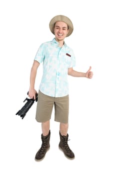 Happy man tourist in blue shirt, brown shorts and hat holds big dslr camera, giving thumbs up and smiles. Traveler going to photo tour. Isolated on white background.
