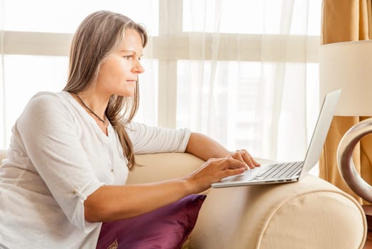 Mature attractive woman is working on laptop while sitting on a couch in her living room