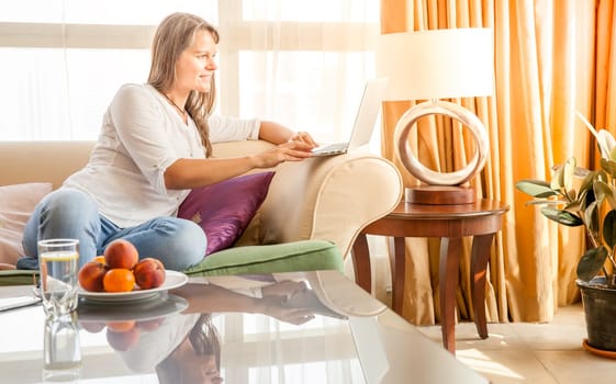 Mature attractive woman is working on laptop while sitting on a couch in her living room