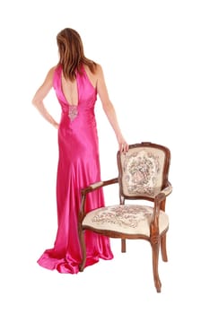 A beautiful young woman in a long pink evening gown standing beside .an old antique armchair from the back, isolated for white background.