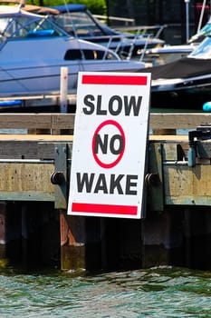 A slow no wake sign on a dock with boats in the background.
