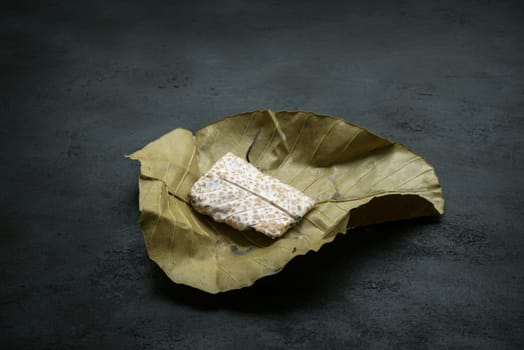 Fresh raw chickpea tempeh wrapped in dillenia suffruticosa leaf, dark background.