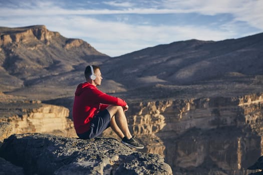 Relaxation in mountains. Young man with headphones sitting on the edge of cliff and listening music. Jebel Akhdar, Grand Canyon of Oman.