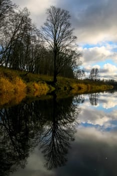 View on autumn landscape of river and trees in sunny day. Forest on river coast in autumn day. Reflection of clouds and trees in water. Autumn in Latvia. Autumn landscape with colorful trees, yellow grass and river.