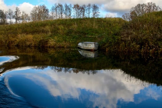 View on autumn landscape of river and boat in sunny day. Forest on river coast in autumn day. Reflection of coast, clouds  and boat in water. Autumn in Latvia. Autumn landscape with trees grass and boat in river.