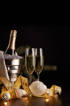 Still life with champagne bottle standing in a bucket with ice, two full champagne flutes, LED lights garland and Christmas ornaments on a black background. Christmas and New Year celebration