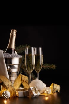 Still life with champagne bottle standing in a bucket with ice, two full champagne flutes, LED lights garland and Christmas ornaments on a black background. Christmas and New Year celebration