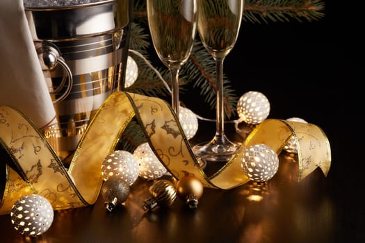 Still life with champagne bottle standing in a bucket with ice, two full champagne flutes, LED lights garland and Christmas ornaments on a black background. Christmas and New Year celebration