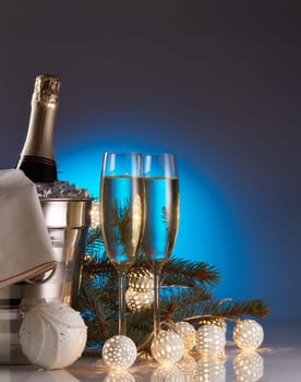Still life with champagne bottle standing in a bucket with ice, two full champagne flutes, LED lights garland and Christmas ornaments. Christmas and New Year celebration background
