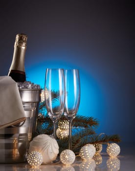 Still life with champagne bottle standing in a bucket with ice, two empty champagne flutes, LED lights garland and Christmas ornaments. Christmas and New Year celebration background