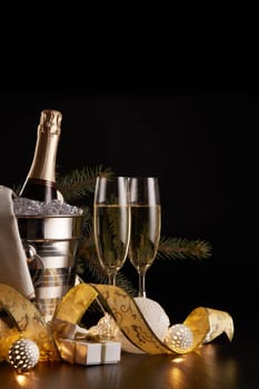 Still life with champagne bottle standing in a bucket with ice, two full champagne flutes, LED lights garland and Christmas ornaments on a black background. Christmas and New Year celebration