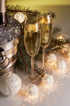 Still life with champagne bottle standing in a bucket with ice, two full champagne flutes, LED lights garland and Christmas ornaments. Christmas and New Year celebration background