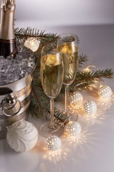 Still life with champagne bottle standing in a bucket with ice, two full champagne flutes, LED lights garland and Christmas ornaments. Christmas and New Year celebration background