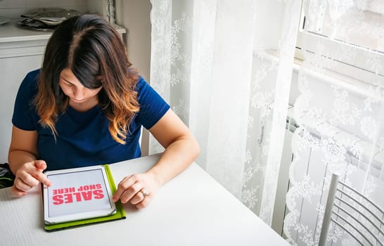 A young brunette woman using a tablet to shop online on a white table with a window and a curtain decorated with white lace. Technology and internet connection concept.