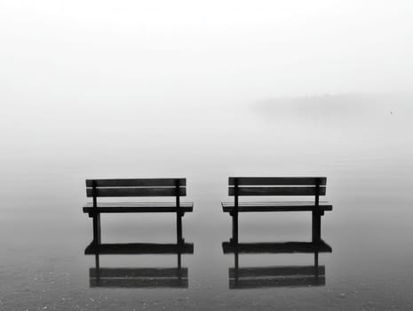 Two empty wooden benches face each other over calm water, shrouded in dense fog. The tranquil setting creates a peaceful atmosphere in the early morning.