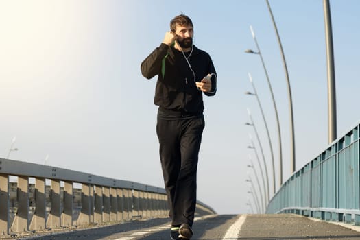 The man in sports clothing is walking on the bridge and listening music with earphones.