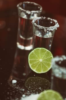 two shots of tequila with lime and salt on a wooden table bar on the background of bright lights of the bar