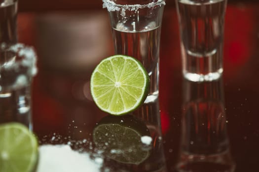 two shots of tequila with lime and salt on a wooden table bar on the background of bright lights of the bar