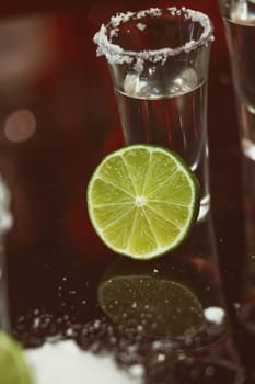 two shots of tequila with lime and salt on a wooden table bar on the background of bright lights of the bar