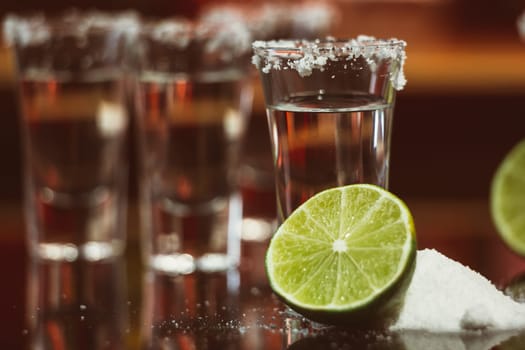 two shots of tequila with lime and salt on a wooden table bar on the background of bright lights of the bar