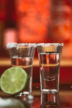 two shots of tequila with lime and salt on a wooden table bar on the background of bright lights of the bar