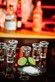 two shots of tequila with lime and salt on a wooden table bar on the background of bright lights of the bar