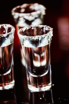 two shots of tequila with lime and salt on a wooden table bar on the background of bright lights of the bar