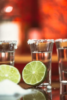 two shots of tequila with lime and salt on a wooden table bar on the background of bright lights of the bar