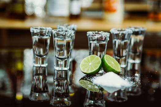 two shots of tequila with lime and salt on a wooden table bar on the background of bright lights of the bar