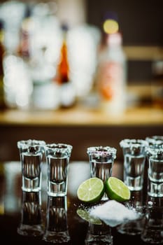 two shots of tequila with lime and salt on a wooden table bar on the background of bright lights of the bar
