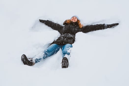Portrait of a beautiful young woman laying down on a frozen snow lake moving her arms and legs up and down creating a snow angel figure, playing games during a sunny winter vacation.