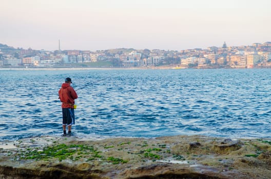 A fisherman in a red rain jacket fishing along the Bondi beach, Sydney, Australia.