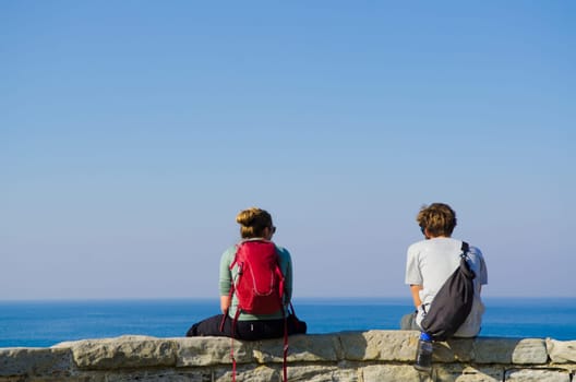 Portrait from behind of young man and woman sitting and looking on Sea with backpack. Concept for travel or freedom.