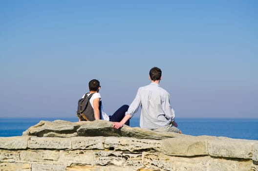 Portrait from behind of young men sitting and looking on Sea with backpack. Concept for travel or freedom.