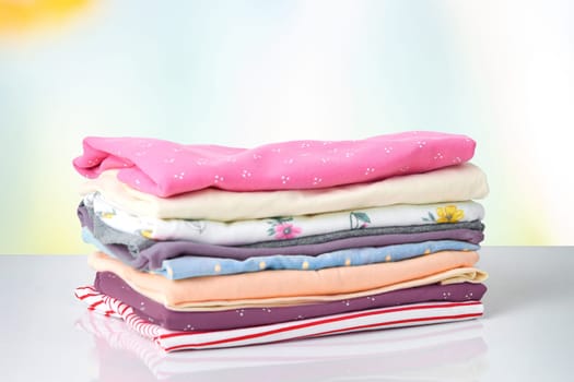 Stack of colorful cute girl clothes. A pile of an colorful clothes on a white table against bright abstract blurred background. Template for your product display montage. Children fashion.