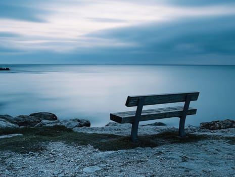 A wooden bench sits on the shore, facing the tranquil sea under a softly lit sky. The calm waters and gentle waves create a peaceful atmosphere during twilight.