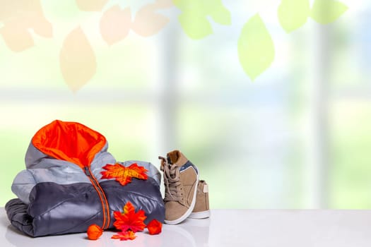 Autumn and winter children clothes. A black gray cozy warm down jacket and a pair shoes with autumn decorations over abstract bright autumn background. Space for your design.