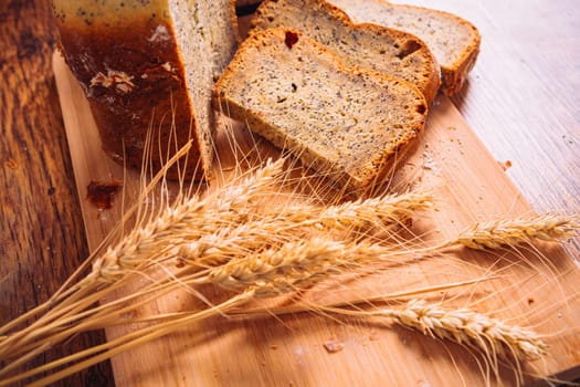 Close slice of fresh bread with poppy seeds and wheat ears on wooden background