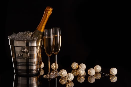 Still life with champagne bottle standing in a bucket with ice, two full champagne flutes, LED lights garland on a black background with copy space. Christmas and New Year celebration