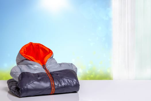 Folded kids colorful autumn jacket. A colorful warm down jacket with orange lining on a white table over abstract light blue sunny autumn day. Template for your product display montage.