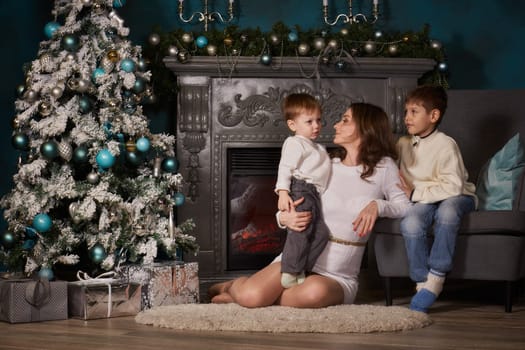 Happy family are sitting near Christmas tree at home. Merry Christmas and Happy New Year. Beautiful young mother with two sons.