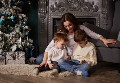 Young mother and her little sons with computer tablet are sitting near Christmas tree at home. Merry Christmas and Happy New Year.
