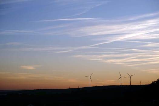 The silhouettes of wind turbines on a wooded hill in the evening..
