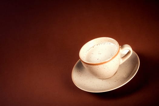 The beige cup of coffee with frothy milk is on dark brown background.