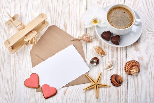 Cup of coffee, cookies and chocolates in the form of heart, seashells, plane and blank sheet with envelope lying on a light wooden background. Top view. Valentine's Day, Honeymoon.