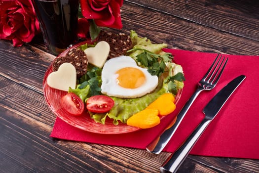 Still life with glass of juice, roses and dish with fried egg, bread, cheese and vegetables in the shape of a heart on a wooden background. Top view. Breakfast on Valentine's Day