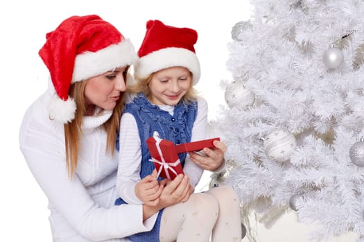 The pretty girl and her mother with gift box are sitting near Christmas tree on a white background.